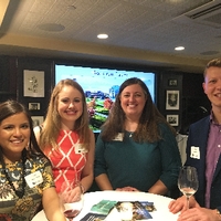 Three young alumni and a parent standing at a table with wine, smiling for camera